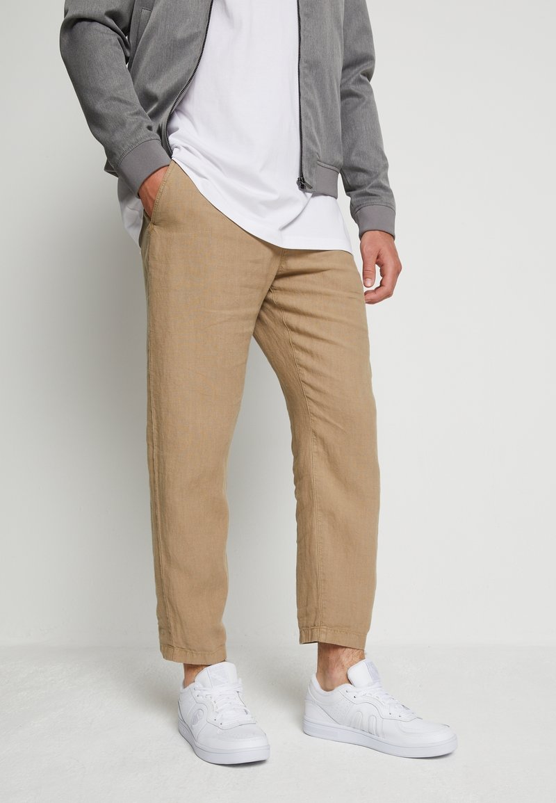 Beige linneblandade byxor med rak skärning och sidofickor, matchade med vita sneakers och en grå bomberjacka över en vit t-shirt.