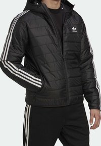 Svart kviltad pufferjacka med huva, med vita tre-stripes accenter på ärmarna och dragkedjefickor på sidan.