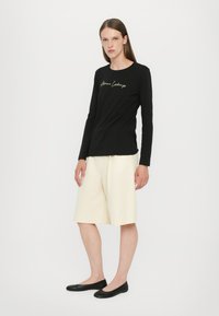 Black long-sleeve shirt with gold "Armani Exchange" text, paired with knee-length cream shorts, and black flat shoes.