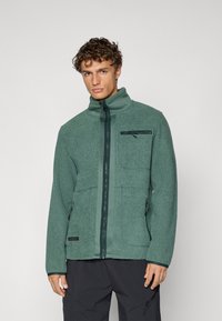Giacca in pile verde con colletto alto, chiusura a zip e due tasche laterali. Presenta una tasca sul petto con zip e tessuto testurizzato.