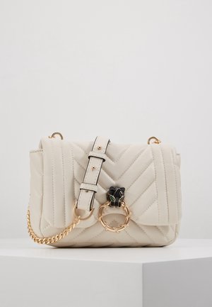 Crèmekleurige doorgestikte crossbodytas met verstelbare band, gouden ketting en zwarte panterkopsluiting met groene ogen die een gouden ring vasthoudt.