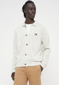 Ανοιχτό γκρι πλεκτή ζακέτα με γιακά, ξύλινα κουμπιά και διακριτή βουτυρωτή υφή. Μικρό ετικέτα λογότυπου στο αριστερό στήθος.