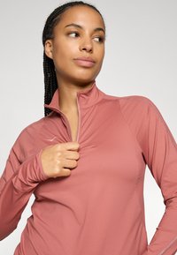 Frau mit geflochtenem Haar, die ein langärmliges rosa Sportoberteil trägt und den Kragen teilweise aufzippt, während sie zur Seite schaut, vor einem neutralen Hintergrund.