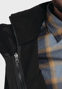 Muž v modro-hnědém kostkovaném tričku pod částečně rozepnutou černou bundou s fleece límcem.