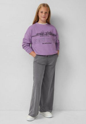 Felpa viola con grafica e testo "NEW YORK", abbinata a pantaloni larghi grigi e sneakers bianche, in piedi davanti a uno sfondo neutro.