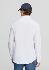 Biała koszula z długim rękawem, z kołnierzykiem zapinanym na guziki, gładka w dotyku, z zaokrąglonym dołem. W zestawie z niebieskimi dżinsami i granatową czapką.