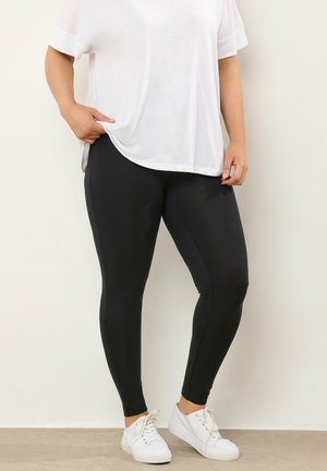 Schwarze Leggings aus elastischem Material mit schlankem Schnitt und glatter Textur, kombiniert mit einem lockeren weißen T-Shirt und weißen Sneakers.