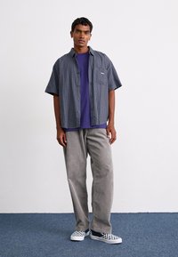 Camisa de cuadros de manga corta en tonos azul y verde, sobre una camiseta morada; combinada con pantalones de pana grises y zapatillas de cuadros sin cordones.