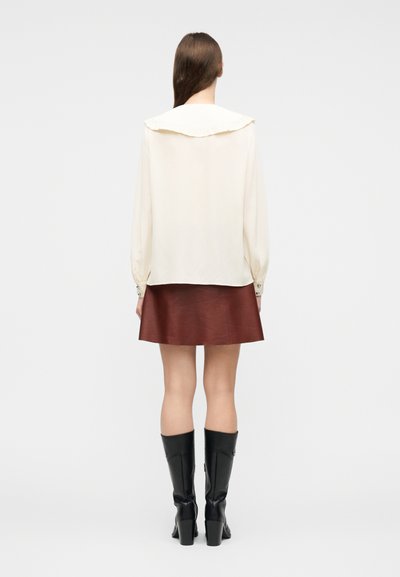 Γυναίκα με μακριά καστανά μαλλιά, που φορά κρεμ μπλούζα με φαρδύ γιακά, καστανή δερμάτινη φούστα και μαύρες μπότες μέχρι το γόνατο, στεκόμενη με την πλάτη προς τα εμάς.