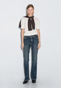 Biała koszula z krótkim rękawem z ciemnym szalikiem zawiązanym na szyi, w połączeniu z dżinsami w stylu dzwonów w kolorze niebieskim i czarnymi butami na szpilkach.