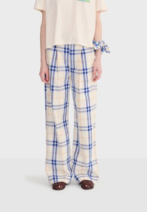 Person iført løstsiddende hvide bukser med blå og orange ternet mønster, brune åbne sandaler og et matchende ternet stof bundet om håndleddet.