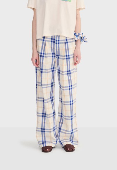 Personne portant un pantalon ample blanc avec un motif à carreaux bleus et oranges, des sandales marron à bouts ouverts, et un tissu à carreaux assorti attaché au poignet.