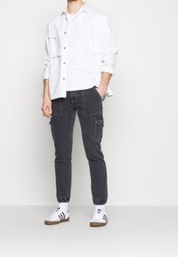 Camisa blanca de botones sobre una camiseta blanca, combinada con pantalones cargo oscuros con bolsillos laterales y zapatillas blancas con rayas negras.