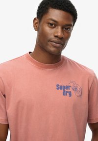 Camiseta de manga corta en un rosa desgastado, hecha de algodón suave. Presenta un texto gráfico en azul que dice "Super dry" y un personaje ilustrado en el pecho.