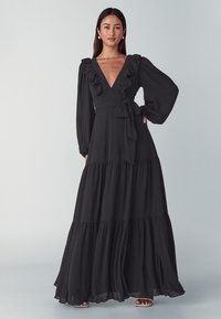 Black maxi dress features a deep V-neckline, ruffled accents on sleeves, a tie waist, and tiered fabric layers for a flowing silhouette.