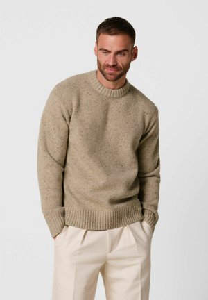 Man met kort haar en baard, gekleed in een beige gebreide trui en crèmekleurige broek, staand met handen in de zakken tegen een witte achtergrond.