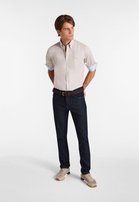 Camisa beige clara de botones con mangas cortas, puños a rayas azules, jeans de mezclilla oscuros, cinturón trenzado marrón y zapatillas beige con acentos amarillos.