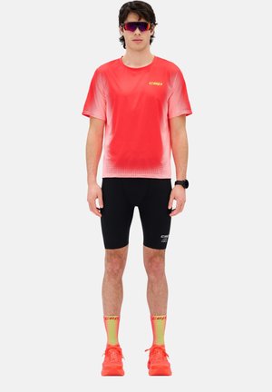 Mann trägt ein rot-weiß verlaufendes Sportshirt, schwarze Kompressionsshorts, neon-gelbe und rote Socken, rote Laufschuhe, Sonnenbrille und eine schwarze Smartwatch.