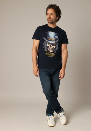 T-shirt marine affichant un graphique de crâne avec un chapeau haut de forme et des accents en spirale, associé à un jean sombre et des baskets blanches avec des détails bleus.