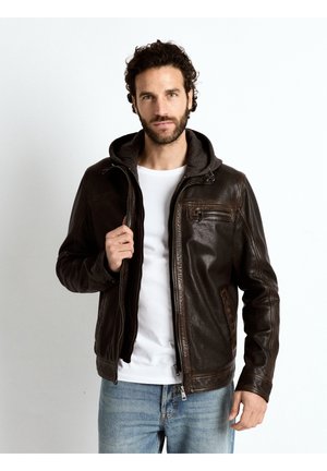 Man draagt een donkerbruine leren jas met capuchon over een wit T-shirt en blauwe spijkerbroek, staande tegen een witte achtergrond.