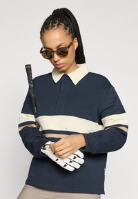 Granatowa koszulka polo z długim rękawem w kremowo-beżowe paski, z gładkim kołnierzem. Model trzyma kij do golfa, ma na dłoniach białe rękawiczki i nosi okulary przeciwsłoneczne.