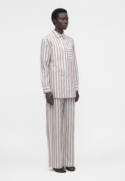 Άτομο που στέκεται, φοράει άνετο πουκάμισο με μπεζ και λευκές κάθετες ρίγες και ταιριαστό παντελόνι με φαρδιά πόδια και μαύρα σανδάλια.
