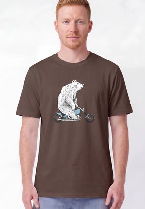Mann, der ein braunes T-Shirt mit einem weißen Bären, der ein blaues Motorrad fährt, auf der Vorderseite trägt, steht vor einem einfarbigen Hintergrund.