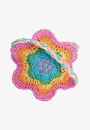 Mladé dievča má na sebe biely bezrukávový odev zdobený farebnými kvetinovými vzormi, háčkovaný slnečný klobúk a viacfarebnú tašku v tvare kvetu.