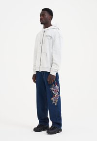 Lys grå zip-up hættejakke med broderet logo, parret med løse mørkeblå jeans med farverigt dragebroderi på venstre ben. Sorte sneakers.