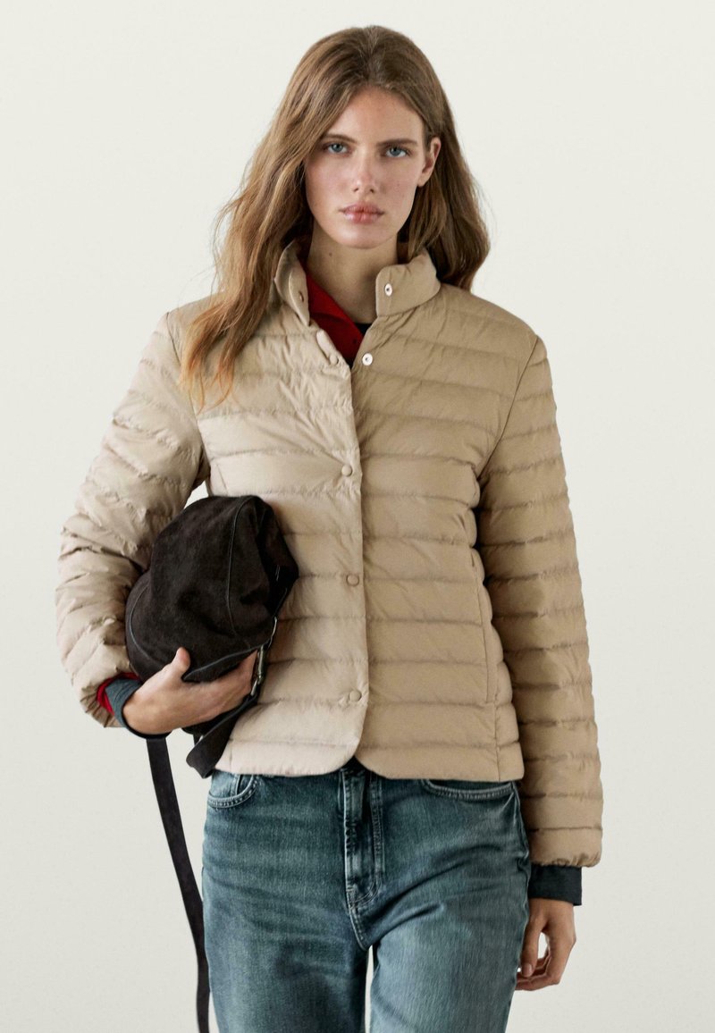 Donna con capelli lunghi che indossa una giacca trapuntata beige e jeans blu, che tiene una morbida borsa a zaino marrone scuro al suo fianco.