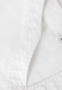 Tela blanca con una correa texturizada que presenta un patrón en zigzag. El fondo es liso y sencillo, lo que resalta los detalles de la correa.