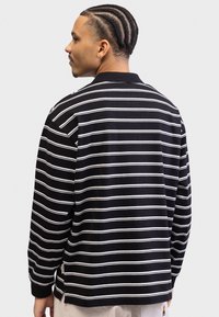 Chemise à manches longues rayée noir et blanc avec un col, présentant une coupe décontractée et des fentes sur les côtés. Matière en coton doux avec des détails de couture visibles.