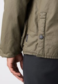 Chaqueta verde oliva con un acabado texturizado, que presenta un bolsillo lateral, cierre de gran botón y una etiqueta a lo largo de la costura. Los pantalones negros son parcialmente visibles.