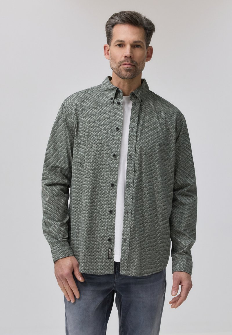 Homme portant une chemise verte à motifs boutonnée sur un t-shirt blanc et un jean bleu délavé, debout devant un fond clair uni.