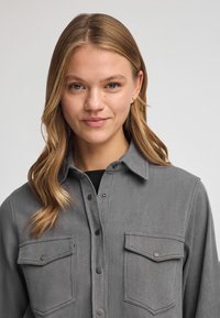 Grijze overhemd met knopen, voorzien van twee borstzakken, een kraag en een zachte textuur. Metalen knopen bieden detail en contrast.