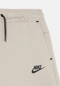 Beige sweatpants with black drawstring, side pockets, and a black Nike logo. Soft fabric with a smooth texture, featuring minimal detailing.