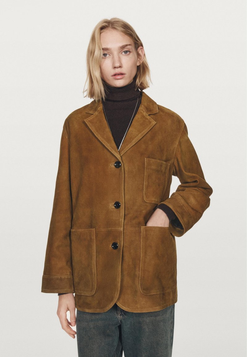 Giubbotto in suede marrone con colletto a pistagna, tre bottoni neri, due tasche frontali e maniche rilassate. Indossato su un dolcevita scuro.