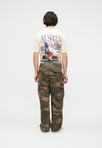 Άτομο που κοιτάζει μακριά, φοράει λευκό T-shirt με κείμενο και γραφικά Austin Texas, παντελόνι καμουφλάζ cargo και λευκά αθλητικά σε απλό φόντο.