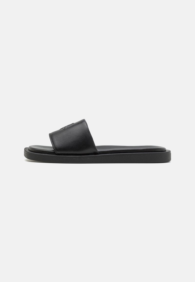 Sandale mule noire avec une large bride en cuir et une semelle plate sur fond blanc, vue de profil.