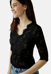 Schwarzes Spitzentop mit einem gewellten V-Ausschnitt und kurzen Ärmeln. Filigrane florale Muster, weiche Textur, kombiniert mit hellblauem Denim.