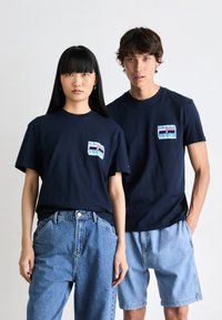 Mørkeblå bomulds T-shirts med korte ærmer, der har et lille flerfarvet mærke på venstre brystområde, iført med denim nederdele.