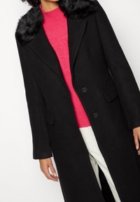 Abrigo largo negro con un collar de pelo sintético, solapas muescas y dos bolsillos frontales, superpuesto a un suéter de punto rosa y pantalones blancos.