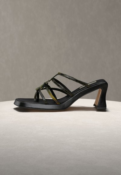 Μαύρο πέδιλο με τακούνι, τετράγωνη μύτη και πολλαπλούς λεπτούς, σκούρους πράσινους ιμάντες σε ουδέτερη επιφάνεια και φόντο.