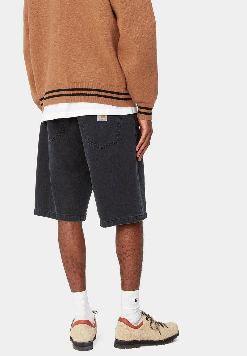 Person iført sorte denimshorts, hvide sokker, beige vandresko, brun sweater med sorte striber, vist bagfra mod en hvid baggrund.