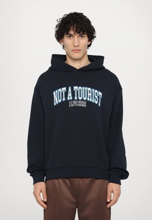 Navy blue hoodie with a front kangaroo pocket, featuring white and light blue text that reads "NOT A TOURIST I LIVE HERE." Soft texture.