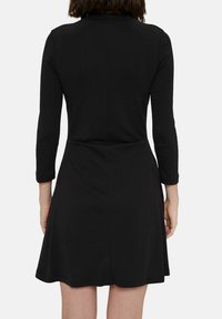 Svart klänning med lång ärm, figursydd överdel och utsvängt nederdel. Slät, elastisk tyg med minimalistisk design. Bakvy visar sömdetaljer.