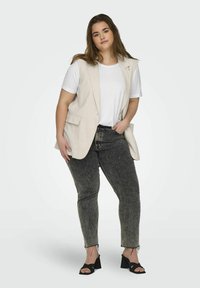 Gilet en lin beige sur un t-shirt blanc, associé à un jean gris foncé. Des sandales à talons noires complètent la tenue. Design simple et minimaliste.
