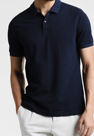 Mężczyzna ubrany w dopasowaną granatową koszulkę polo z krótkim rękawem i jasne spodnie, stojący z jedną ręką w kieszeni.