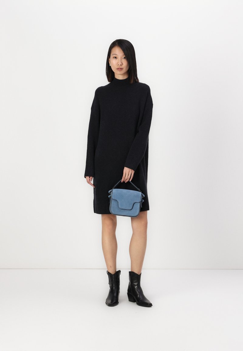 Schwarzes Strickkleid mit hohem Ausschnitt, kombiniert mit einer kleinen blauen Wildleder-Handtasche und schwarzen Stiefeletten. Glatte Textur und strukturiertes Design.