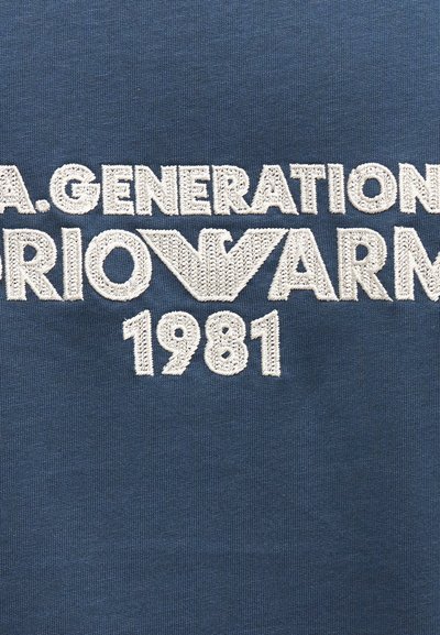 Μπλε ύφασμα με λευκό κεντημένο κείμενο που γράφει "A.GENERATION" πάνω από το "ARMANI" με ένα στυλιζαρισμένο λογότυπο "A" και το έτος "1981" από κάτω.